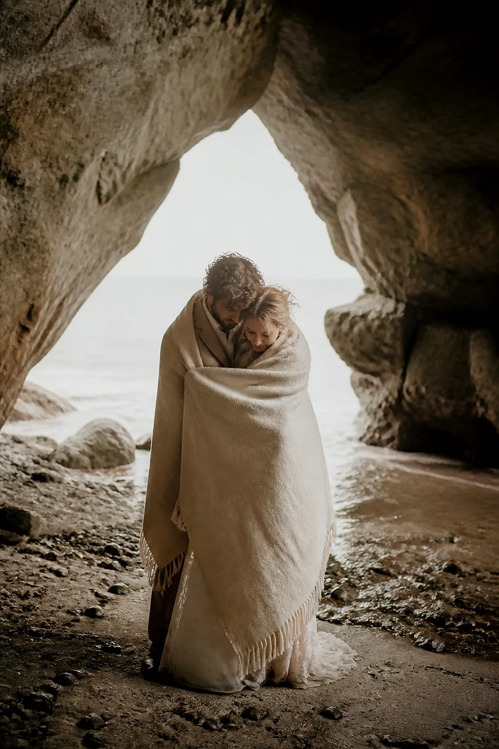 An eloping couple wrapped in a wool blanket at the mouth of a sea cave